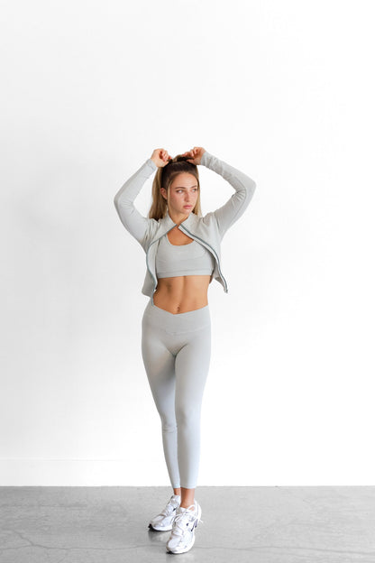 Model with arms raised in the sage SYLA Sculpt Set, cropped jacket worn open over bra and leggings